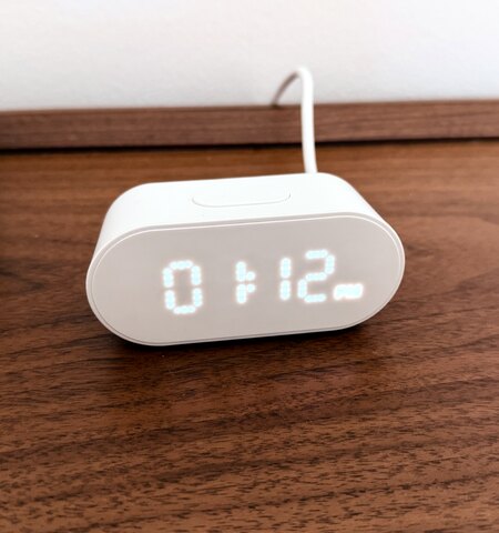 An IKEA ALPSTUGA on a wood desk showing 1:12 PM on its LED dot-matrix display
